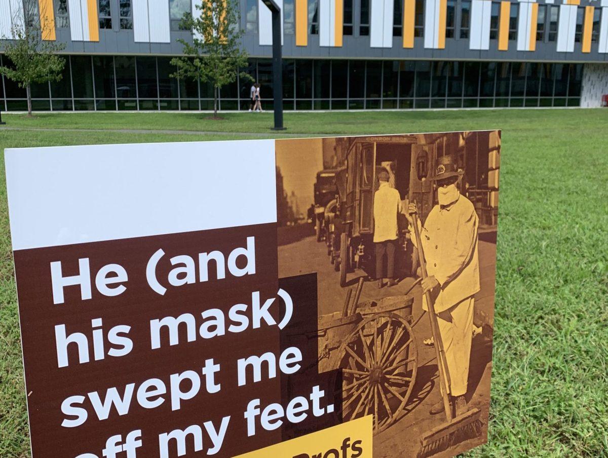 A sign reading "He (and his mask) swept me off my feet" and featuring a vintage photograph of a couple stands outside of Holly Pointe Commons residence hall. This sign is one of many that have been placed across campus by Lori Marshall and the University Publications team to keep the Rowan community aware of pandemic-era public health protocols. - Image courtesy of University Publications.